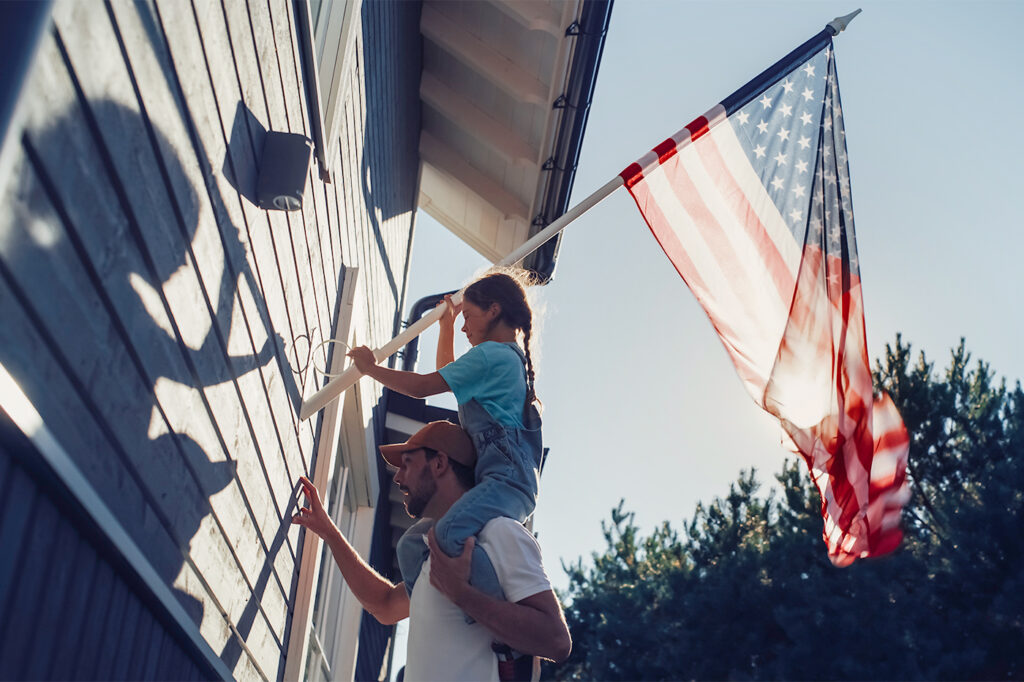 Cinematic Shot of a Father Holding His Small Daughter of His Shoulders, Helping Her to Raise the United States of America Flag to Celebrate a National Fourth of July Holiday at Their House.